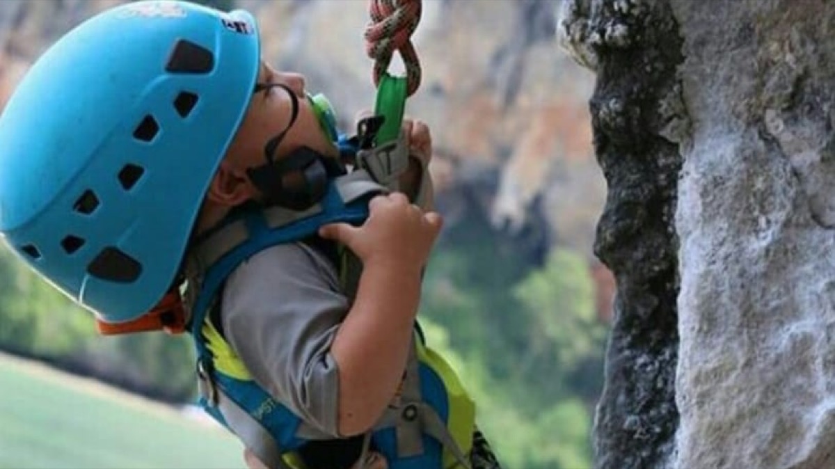 A young child in a blue helmet and harness clipping into a rope on a rock face